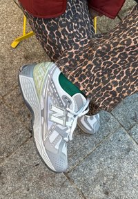 Pilki New Balance sportbačiai turi tinklinę ir sintetinę medžiagą, siuvinėtus akcentus ir raštuotą pado išorinę dalį, kartu dėvimi leopardų rašto kelnės.