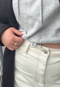 Hall hallatud kapuutsiga dressipluus kootud nööriga ja kõrge vöökohaga kreemikarva lühikeste pükstega, mis on kaunistatud nööpide ja õmbluste detailidega. Kullast sõrmus käel.