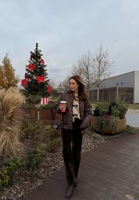 Brązowa pikowana kurtka, beżowy sweter, czarne aksamitne spodnie, czarna torebka. Scena na zewnątrz z udekorowaną choinką i zielenią.