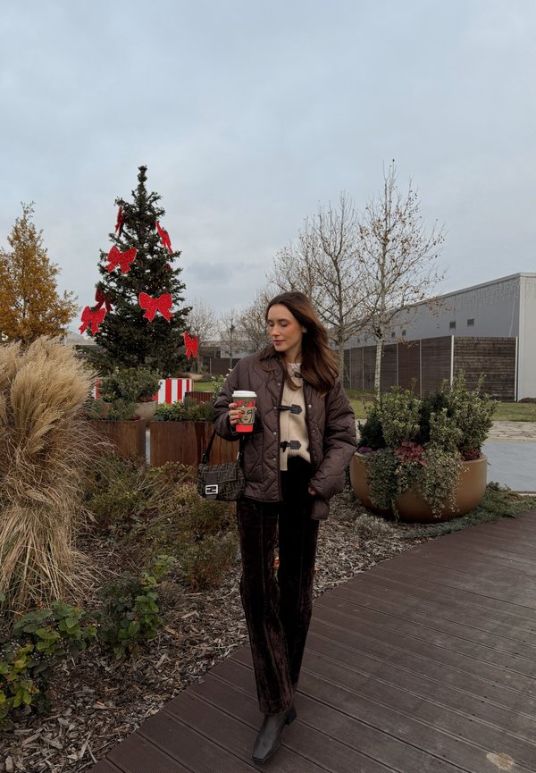 Giacca marrone trapuntata, maglione beige, pantaloni in velluto nero, borsa nera. Scena all'aperto con un albero di Natale decorato e vegetazione.
