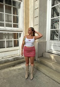 Top corto bianco con grafica rossa, mini gonna a righe rosse e bianche, stivali al ginocchio beige e borsa intrecciata con un dettaglio verde.