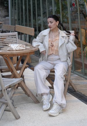 Chaqueta de cuero gris claro sobre una blusa de melocotón con volantes, combinada con pantalones beige y zapatillas blancas. Accesorios de collares de perlas en capas.