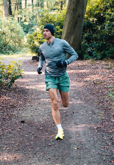 Hombre atlético corriendo por un sendero de tierra, vistiendo una camiseta de manga larga gris, pantalones cortos verdes, guantes negros y zapatillas de correr amarillas.