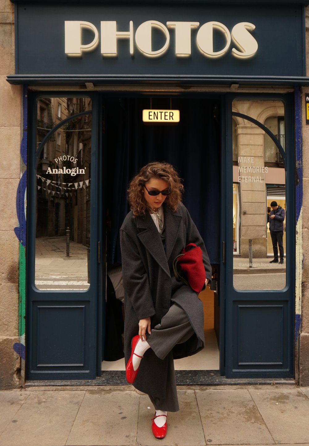 Manteau gris, sac à main rouge, chaussures rouges et chaussettes blanches. La devanture du magasin affiche une enseigne "PHOTOS" ; des rideaux sombres sont visibles à l'intérieur.