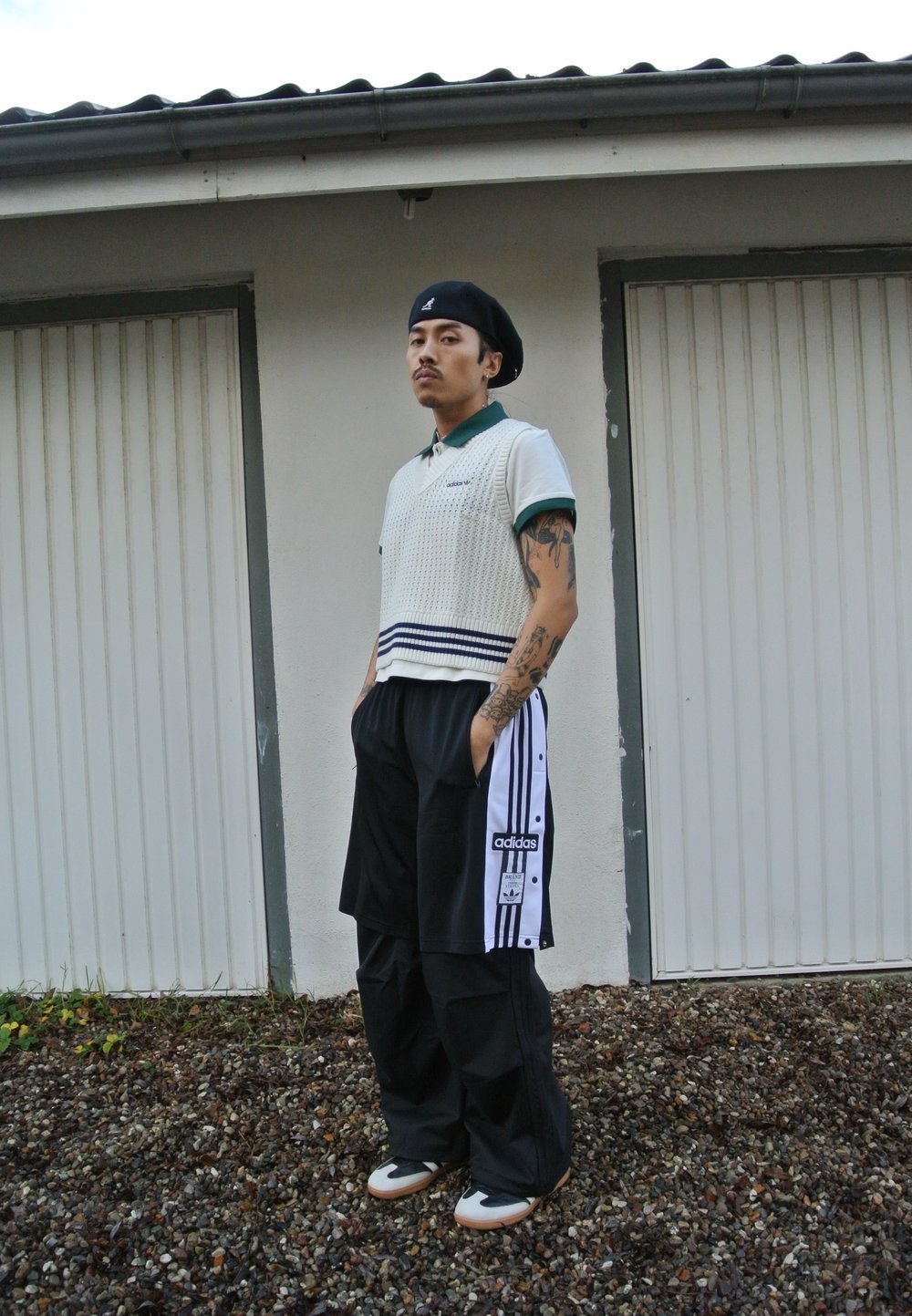 Λευκό πλεκτό πόλο με πράσινη κολόντα, οριζόντιες ρίγες, μαύρο παντελόνι φόρμας με λευκές ρίγες, κρέμα και μαύρα αθλητικά παπούτσια, να στέκεται μπροστά σε έναν τοίχο.