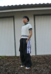 Λευκό πλεκτό πόλο με πράσινη κολόντα, οριζόντιες ρίγες, μαύρο παντελόνι φόρμας με λευκές ρίγες, κρέμα και μαύρα αθλητικά παπούτσια, να στέκεται μπροστά σε έναν τοίχο.