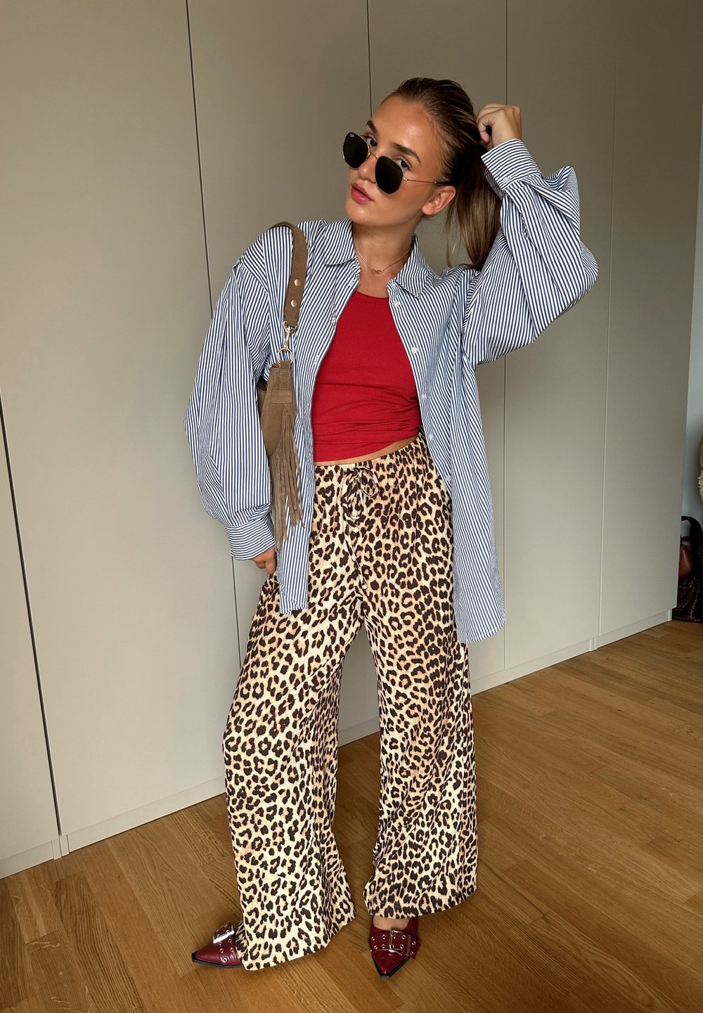 Prugasta košulja na dugih rukava, crni top, široke hlače s leopard uzorkom, crne sunčane naočale i crvene štikle u zatvorenom prostoru.