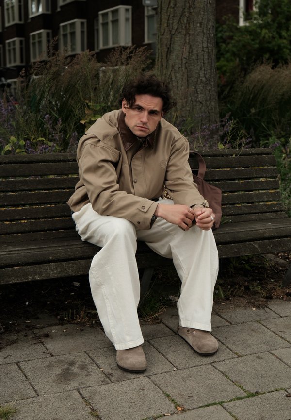 Mann trägt beige Jacke und weiße Hose, sitzt auf einer Holzbank im Freien mit Pflanzen und einem Baum hinter sich.