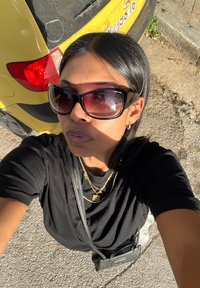 Μαύρο T-shirt, χρυσά κοσμήματα, υπερμεγέθη μαύρα γυαλιά ηλίου, σκούρα μαλλιά, μαύρη τσάντα. Κίτρινο αυτοκίνητο στο βάθος κάτω από το ηλιακό φως, ρίχνοντας σκιές.