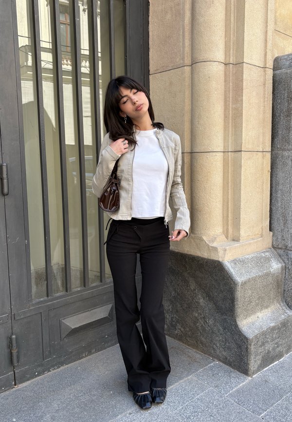 Femme aux yeux fermés debout près d'une grande fenêtre et d'un mur en pierre, portant un haut blanc, une veste beige, un pantalon noir, tenant un petit sac à main marron.