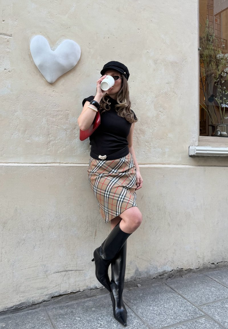 Femme portant une casquette noire, un débardeur, une jupe à carreaux et des bottes noires montantes, buvant dans une tasse blanche tout en s'appuyant contre un mur avec un cœur blanc.