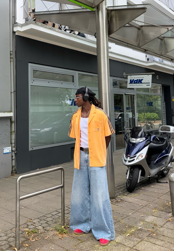 Orange skjorta med svarta ränder, vit linne, vida blå denimbyxor och röda sneakers. Bär hörlurar och står vid en metallstång.