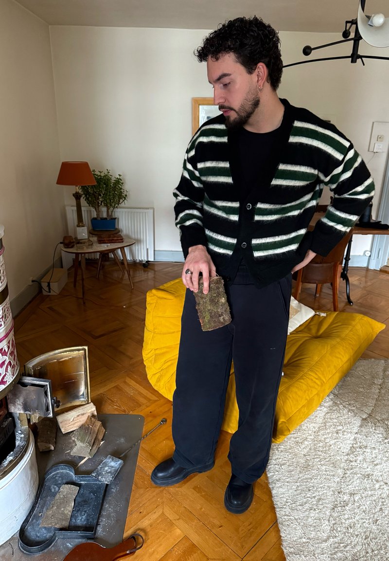 Gestreifte schwarz-grüne Strickjacke, schwarzes Hemd und Hose; hält ein kleines, mit Moos bedecktes Holzstück neben einem Holzofen, im Hintergrund ein gelbes Sofa.