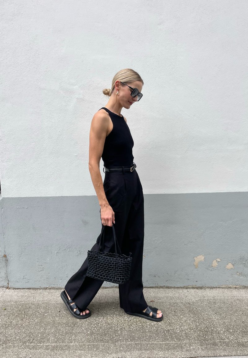 Top senza maniche nero, pantaloni neri a vita alta e larghi, sandali neri con platform e una borsa a tracolla nera intrecciata con manici corti, camminando su un marciapiede.