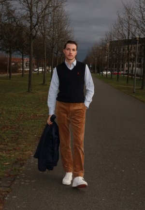 Conjunto de moda que incluye un chaleco de punto azul marino, camisa blanca, pantalones de pana beige y zapatillas blancas. Sosteniendo una chaqueta azul marino.