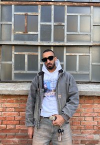 Uomo con occhiali da sole, giacca grigia, felpa bianca con grafica e jeans si trova contro un muro di mattoni e una finestra in metallo ghiacciato tenendo le chiavi.