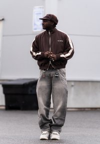 Homme en veste brune zippée avec des rayures blanches, jean gris ample, baskets blanches et casquette brune, debout dehors sur un trottoir.