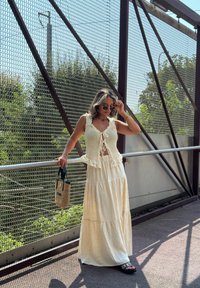 Maglia bianca a maniche corte con pizzo, abbinata a una gonna maxi crema ampia. Accessori includono una borsa intrecciata, indossata con sandali neri.