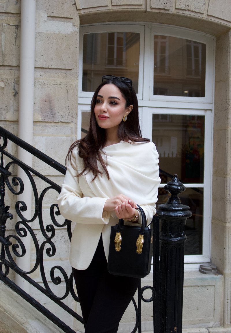 Femme aux longs cheveux foncés portant un blazer blanc et un pantalon noir, tenant un sac à main noir avec des détails dorés, debout près d'une rambarde en fer forgé.