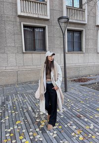 Langer beiger Mantel über einem braunen Crop-Top, schwarzen weit geschnittenen Hosen und beigen Stiefeln. Weißer Hut, lange dunkle Haare und ein leichter Schal. Herbstblätter auf dem Gehweg.