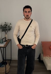 Langärmliges cremefarbenes Ripp-Shirt mit kleinem Logo, kombiniert mit dunkelblauen Jeans. Eine schwarze Umhängetasche und eine strukturierte orangefarbene Decke sind sichtbar.