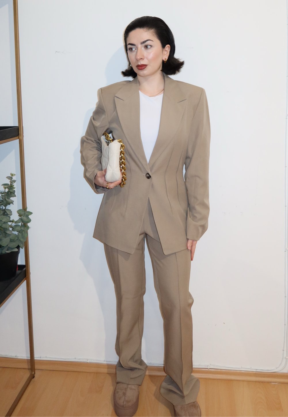 Femme en costume beige tenant un sac à main blanc matelassé avec chaîne dorée, debout à l'intérieur contre un mur blanc avec un sol en bois.