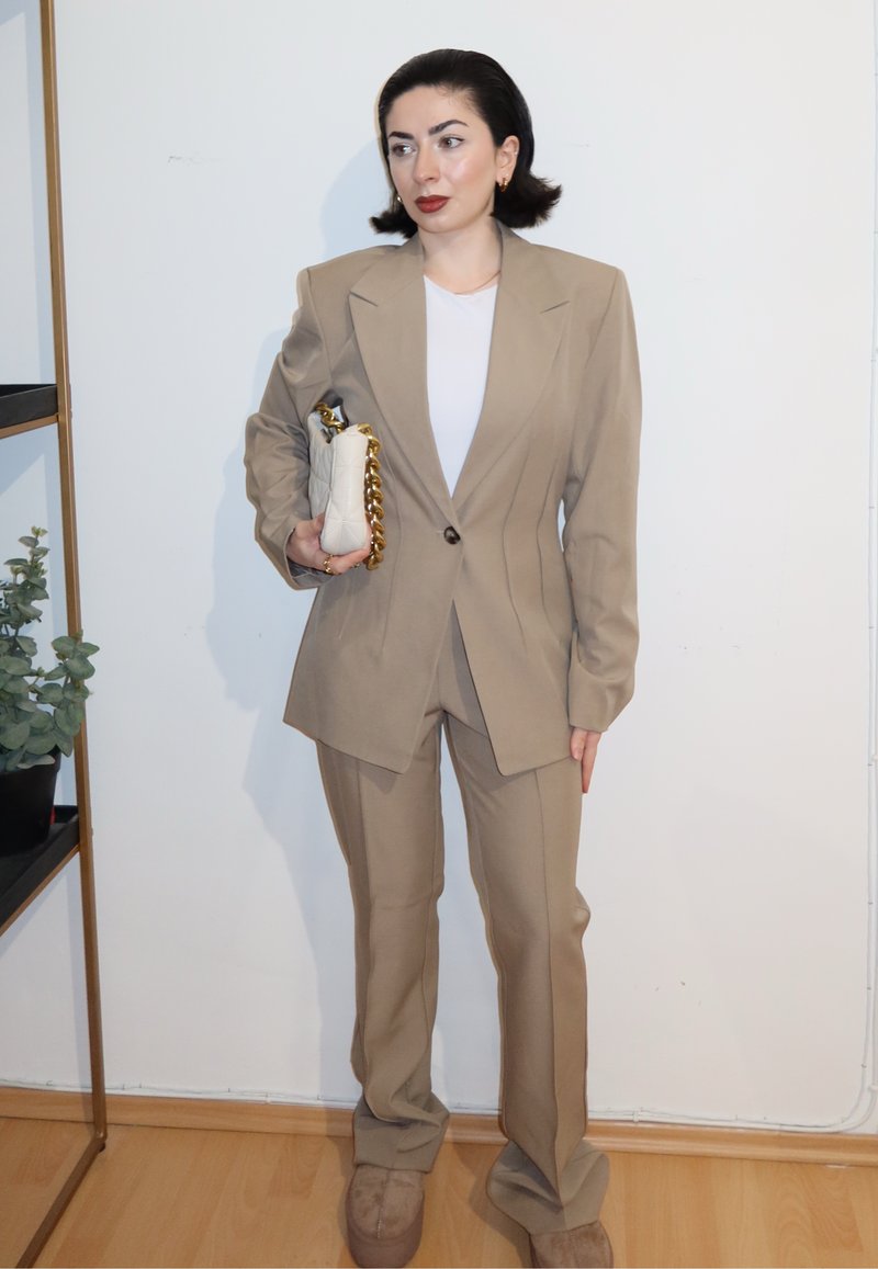 Femme en costume beige tenant un sac à main blanc matelassé avec chaîne dorée, debout à l'intérieur contre un mur blanc avec un sol en bois.