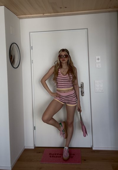 Mujer con cabello largo vistiendo una camiseta sin mangas a rayas rosas y pantalones cortos, zapatillas rosas, gafas de sol, sosteniendo un bolso rosa, de pie frente a una puerta blanca.