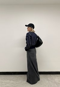 Laivaston raidallinen neule, korkeavyötäröinen harmaa farkku-maksihame, musta lippalakki ja pieni musta laukku, aseteltuna tasaisen seinän eteen.