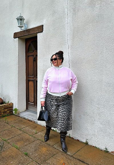 Chaqueta rosa con cremallera y rayas blancas, falda de estampado de leopardo, bolso negro y botas marrones hasta la rodilla contra una pared texturizada.