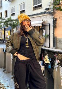 Zelená bomber bunda cez čierny top, čierne tepláky a žltý čiapkový klobúk so vzorom. Príslušenstvo zahŕňa viacero zlatých náhrdelníkov a prsteňov.