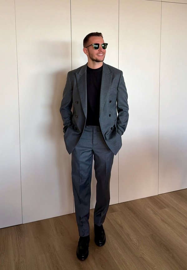 Hombre con gafas de sol oscuras, traje gris cruzado, camisa negra, zapatos negros, de pie en el interior contra una pared blanca y suelo de madera.