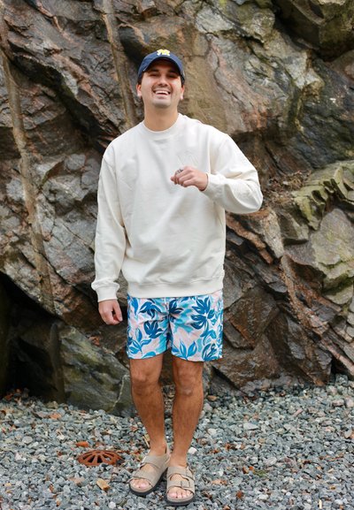 Hombre sonriente con gorra azul, sudadera blanca, pantalones cortos florales y sandalias, de pie sobre terreno rocoso frente a una gran pared de piedra rugosa.