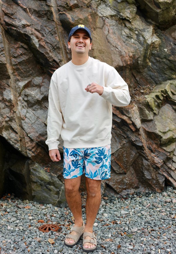 Glijdende man met een blauwe pet, witte sweatshirt, bloemige korte broek en sandalen staat op rotsachtige grond voor een grote ruwe stenen muur.