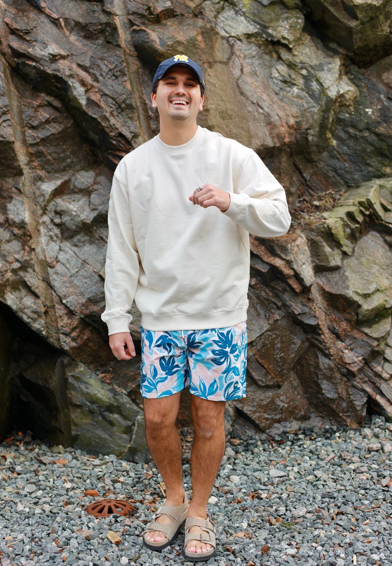 Lächelnder Mann mit blauer Kappe, weißem Sweatshirt, floralen Shorts und Sandalen steht auf felsigem Boden vor einer großen rauen Steinmauer.
