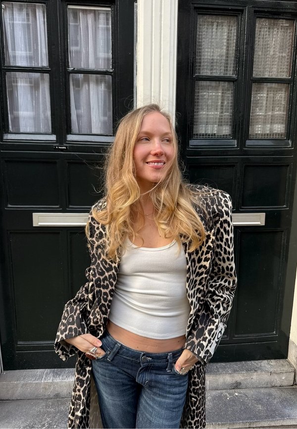 Jeune femme portant un manteau à imprimé léopard, un débardeur blanc et un jean, debout en souriant, les mains dans les poches, devant une porte noire.