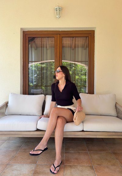Mujer con gafas de sol, vistiendo un top negro y pantalones cortos, sentada en un sofá exterior beige acolchado sosteniendo una pequeña bolsa tejida, con una ventana de madera detrás.