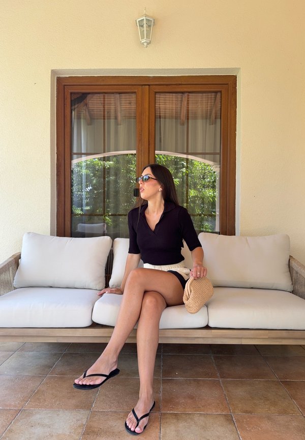 Mujer con gafas de sol, vistiendo un top negro y pantalones cortos, sentada en un sofá exterior beige acolchado sosteniendo una pequeña bolsa tejida, con una ventana de madera detrás.