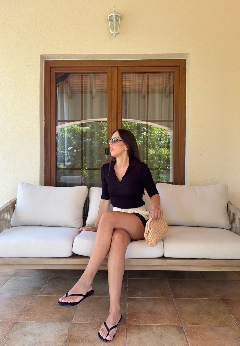 Femme portant des lunettes de soleil, un haut noir et un short, assise sur un canapé d'extérieur beige rembourré, tenant un petit sac tissé, avec une fenêtre en bois derrière elle.