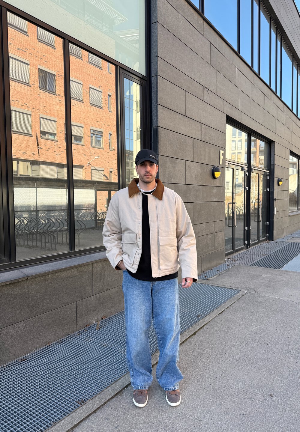 Beige polstret jakke med brun krage, sort genser, løse blå jeans og grå joggesko. Står nær en moderne bygning med store vinduer.