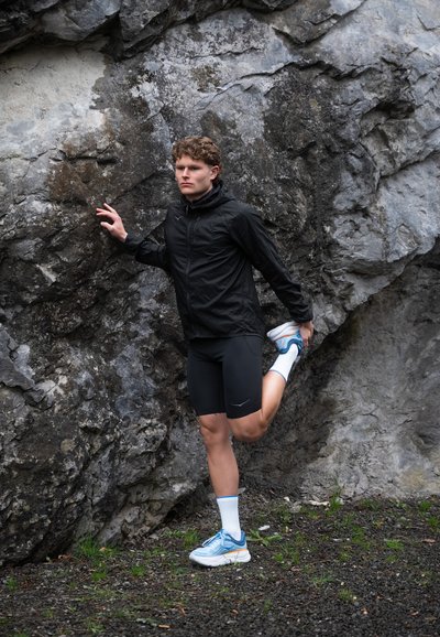 Chaqueta ligera negra, pantalones cortos negros, zapatillas de correr azules con suelas blancas y calcetines hasta el tobillo. Persona estirándose contra un fondo rocoso.