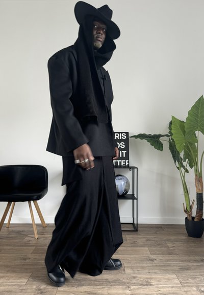 Conjunto negro de capas que incluye un abrigo largo, pantalones anchos y un sombrero grande. Textura suave con detalles visibles de costuras. Los accesorios incluyen anillos.