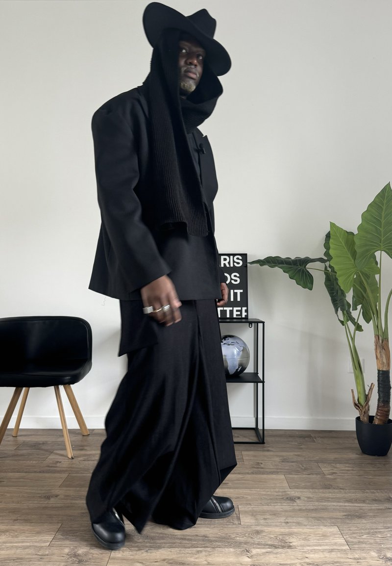 Black layered outfit featuring a long coat, wide pants, and a large hat. Smooth texture with visible stitching details. Accessories include rings.