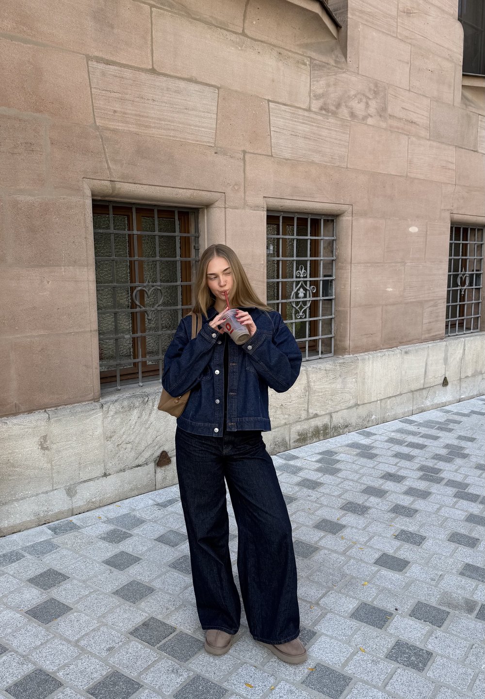 Donkerdenim jas met knoopsluiting, gecombineerd met wijde donkere jeans. Houdt een drankje met een rietje vast; beige tas over haar schouder.