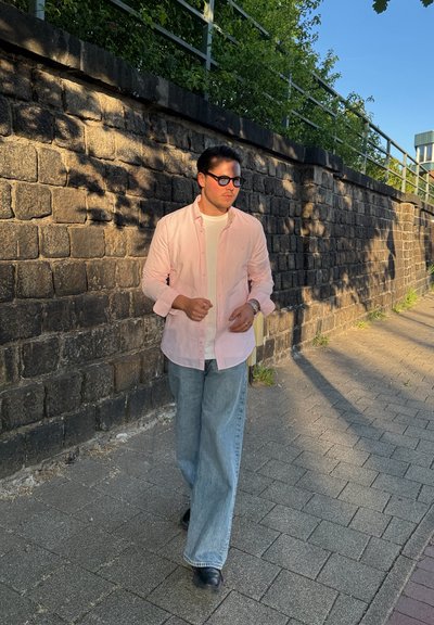 Camisa de manga larga rosa claro, superpuesta a una camiseta blanca, combinada con jeans de mezclilla holgados y zapatos negros. Muro de piedra texturizado de fondo.