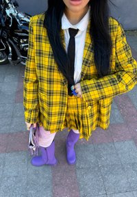 Blazer oversized amarelo xadrez e saia combinando, camisa branca, gravata preta, botas roxas até ao joelho, e a segurar uma clutch prateada.