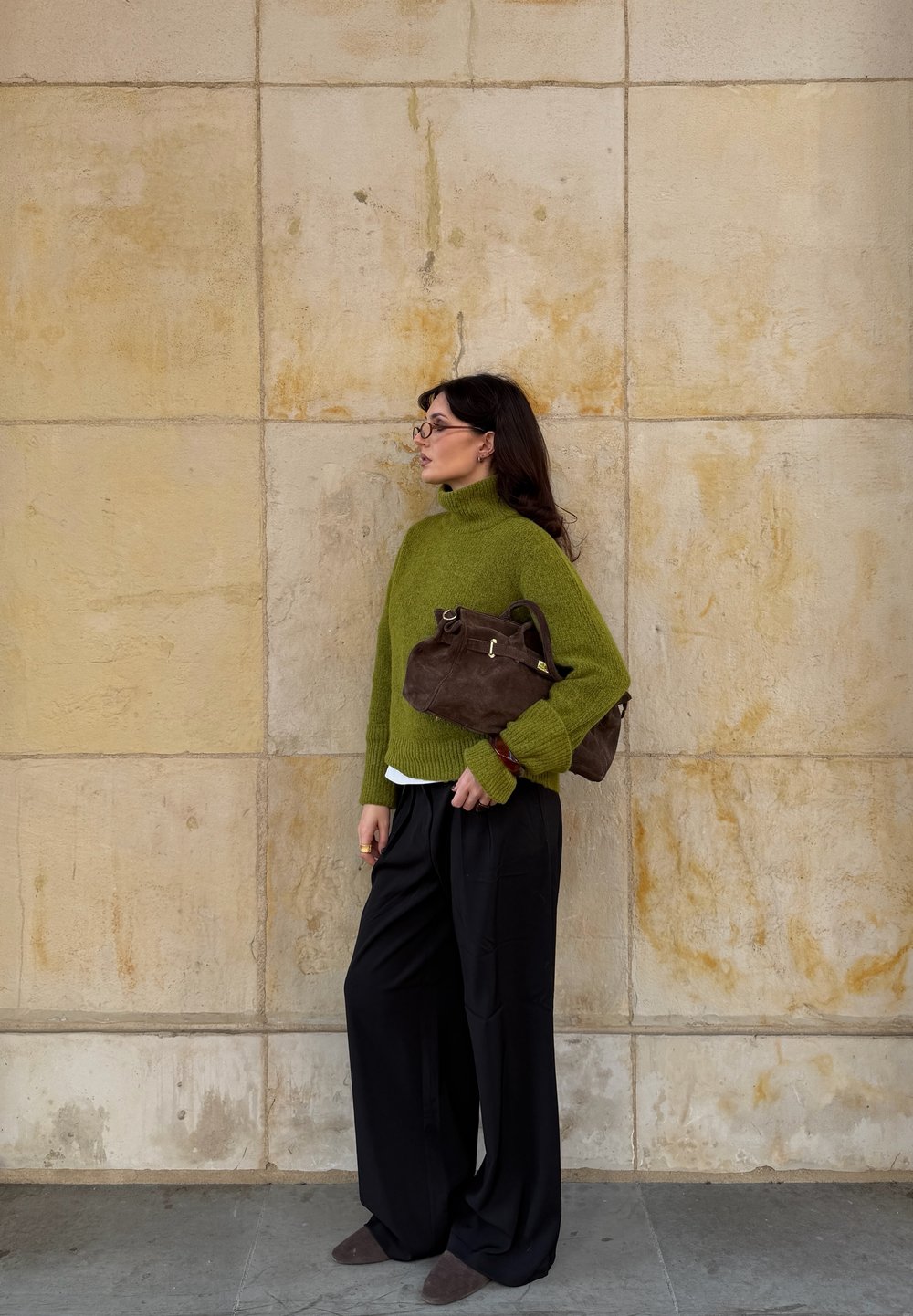 Grøn strikket turtleneck sweater, løstsiddende sorte bukser, brun ruskinds håndtaske, stående mod en struktureret lys væg.