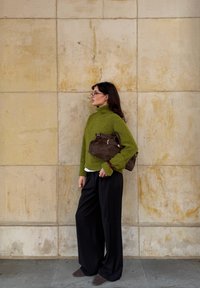 Grøn strikket turtleneck sweater, løstsiddende sorte bukser, brun ruskinds håndtaske, stående mod en struktureret lys væg.