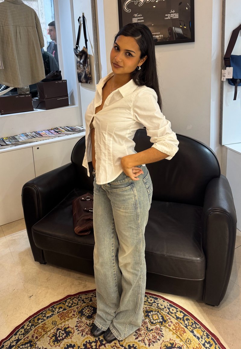 Femme aux longs cheveux foncés se tient la main sur la hanche, portant un chemisier blanc et un jean bleu clair dans une pièce cosy avec un canapé en cuir noir et un tapis à motifs.