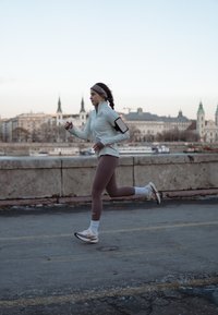 Tenue de course : veste zippée gris clair, leggings marron, chaussettes de sport blanches et baskets avec des touches rose pâle. Paysage urbain en arrière-plan.